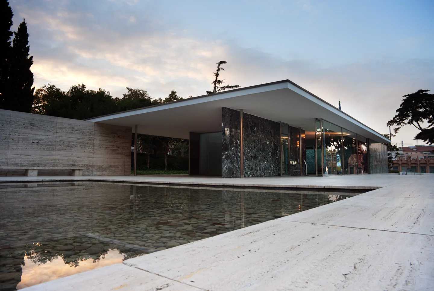 Pavilhão de Barcelona ao entardecer: planos de pedra e vidro sob uma cobertura horizontal, junto a um espelho d’água.