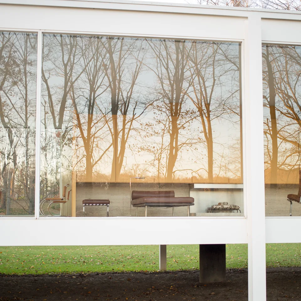 Fachada envidraçada da Casa Farnsworth, com estrutura branca e árvores refletidas no vidro.